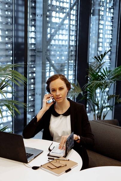 Business portrait. The business woman in a white blouse and a black jacket talks on the smartphone in a workplace at office. Nearby on a table a latte cup of coffee, a dessert, a notebook and office supplies. - Foto, Imagem