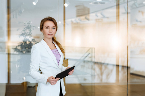 Business muotokuva positiivinen nainen blondi valkoisessa takissa taustaa vasten modernin toimiston business center musta tabletti käsissä
. - Valokuva, kuva