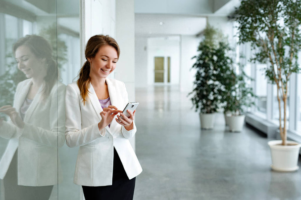 Obchodní a portrét pozitivní ženy blondýna v bílém plášti na pozadí moderní kancelář obchodního centra. Děvče hovoří na smartphone. - Fotografie, Obrázek