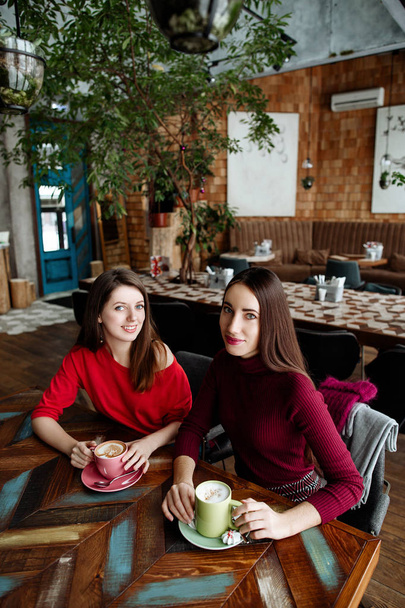 Meeting of girlfriends in cafe. Two fashionable modern girls in red dresses sit and communicate behind a cappuccino cup of coffee. - Foto, afbeelding