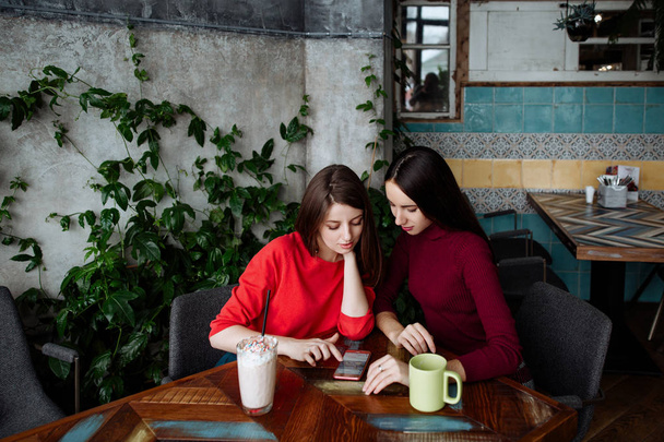 Meeting of girlfriends in cafe. Two fashionable modern girls in red dresses sit and communicate behind a cappuccino cup of coffee. - Foto, imagen