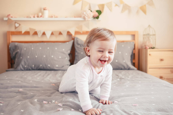 Portrait of cute adorable smiling Caucasian blonde baby girl in white onesie sitting on bed in bedroom decorated for birthday event celebration. Happy childhood lifestyle concept - Zdjęcie, obraz