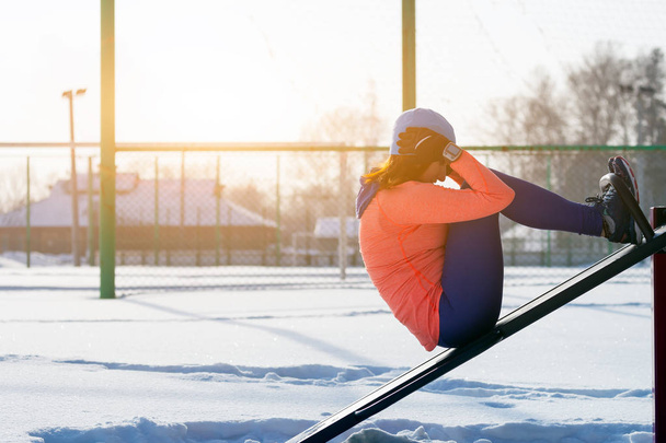 Uma jovem mulher em um chapéu azul brilhante, camisola laranja e espetos de alce pressiona uma prensa em um simulador vertical em um campo de esportes em um dia de inverno brilhante.
 - Foto, Imagem