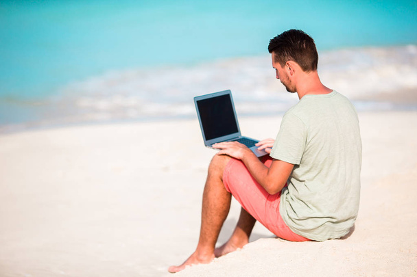 Młody mężczyzna siedzący na piasku z laptopa na tropikalnej plaży Karaibów. Człowiek z komputera i pracy na plaży - Zdjęcie, obraz