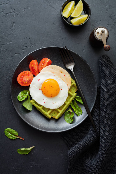 Fried egg with cheese, tomatoes, chard, and waffle with spinach on black ceramic plate on dark concrete background. Sandwich for breakfast. Selective focus. Top view. Copy space. - Foto, immagini