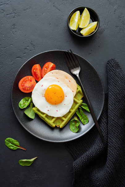 Fried egg with cheese, tomatoes, chard, and waffle with spinach on black ceramic plate on dark concrete background. Sandwich for breakfast. Selective focus. Top view. Copy space. - Фото, изображение