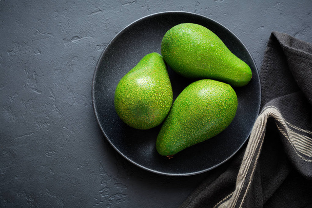 Three raw fresh avocados in black ceramic plate on black concrete background. Selective focus. Top view. - Foto, immagini