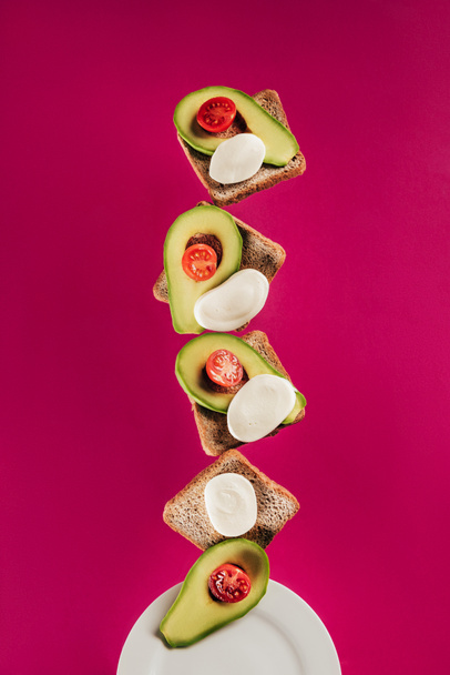 close up view of toasts, avocado pieces, mozzarella cheese and cherry tomatoes falling on plate isolated on pink - Fotografie, Obrázek