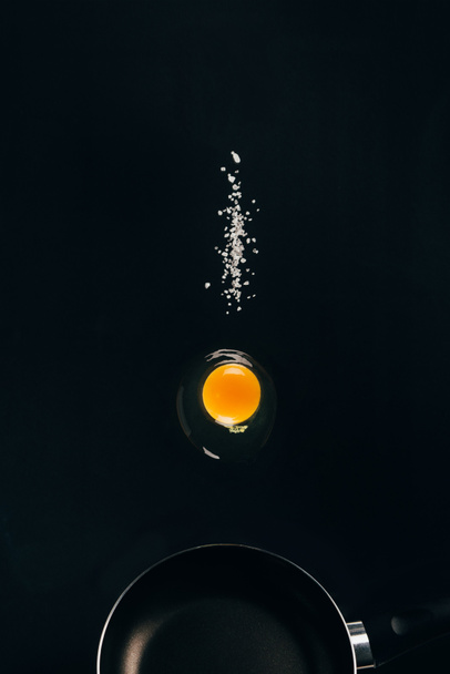 close up view of raw egg yolk and salt falling on frying pan isolated on black - Zdjęcie, obraz