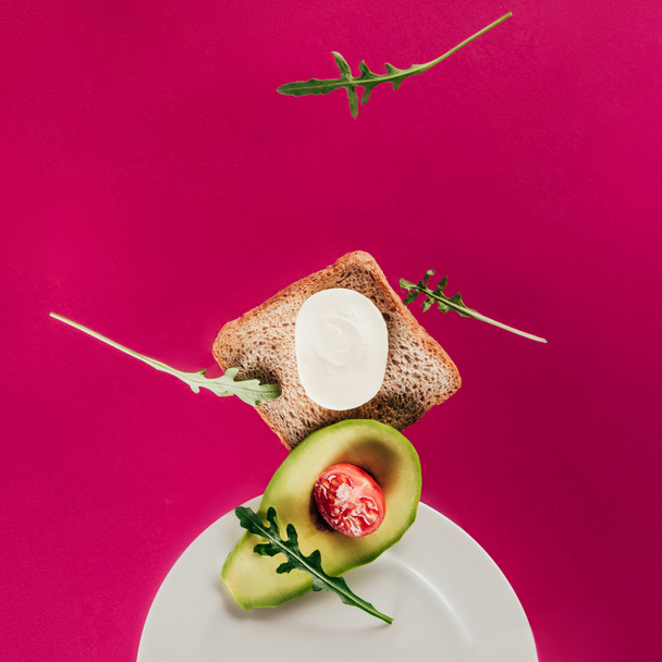close up view of toast, pieces of avocado, cherry tomato, mozzarella cheese and arugula falling on plate isolated on pink - Photo, Image