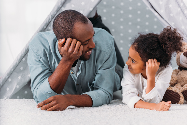 african american father and daughter lying on floor and looking at each other at home - Foto, imagen