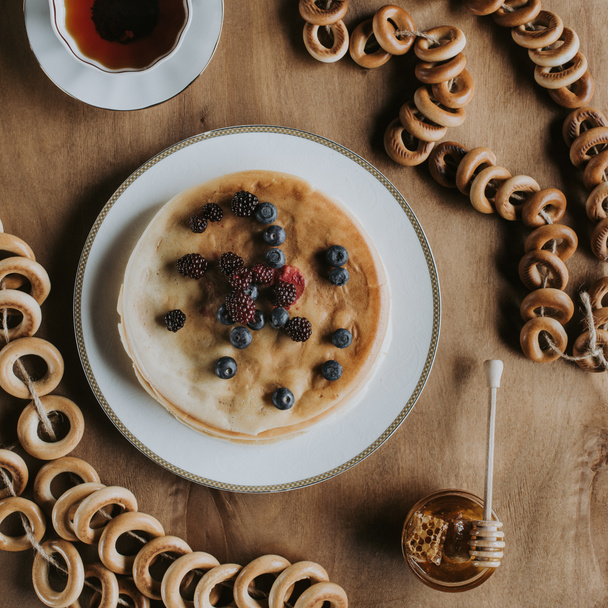 top view of sweet tasty pancakes with berries, cup of tea, honey and bagels on wooden table    - Fotoğraf, Görsel