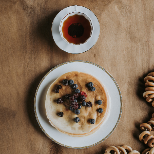 top view of delicious pancakes with berries, cup of tea and bagels on wooden table   - Foto, afbeelding