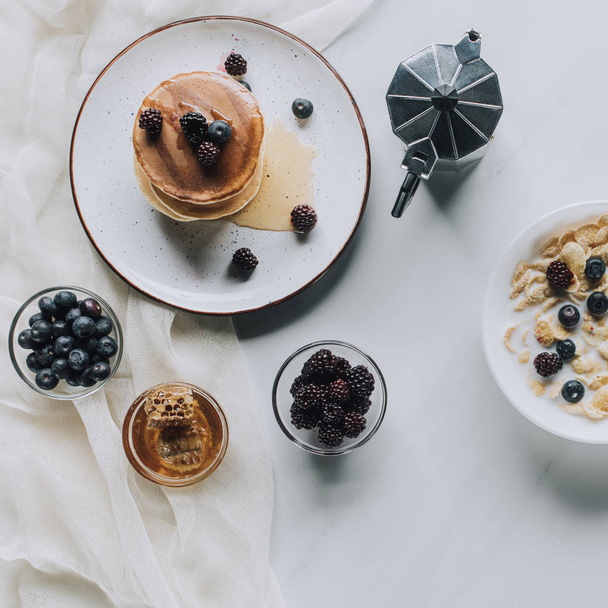 top view of fresh homemade pancakes with honey and berries, coffee pot and muesli with milk on grey - 写真・画像