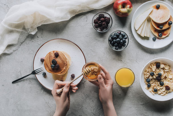 bovenaanzicht van persoon eten heerlijke huisgemaakte pannenkoeken met honing en fruit op grijs - Foto, afbeelding