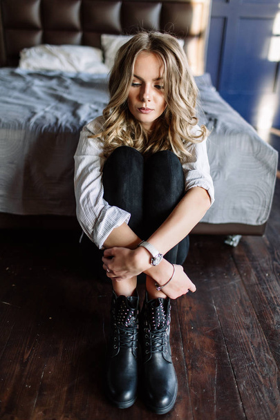 A girl with long blond hair dressed in black jeans, a striped shirt and black shoes sitting on the bed with blue linens. Fashionable casual outfit. - Fotoğraf, Görsel