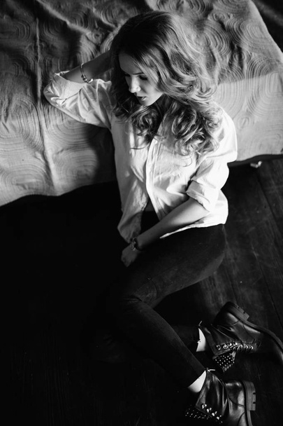 Een meisje met lang blond haar, gekleed in een gestreepte shirt, zwarte jeans en zwarte schoenen zitten op het bed met blauwe beddengoed. Modieuze casual outfit. - Foto, afbeelding
