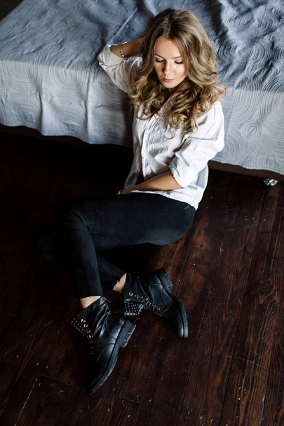 A girl with long blond hair dressed in black jeans, a striped shirt and black shoes sitting on the bed with blue linens. Fashionable casual outfit. - Foto, Imagen