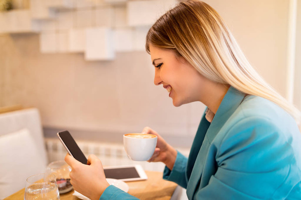 Молодая улыбающаяся деловая женщина на перерыве в кафе. Он пьет кофе и читает сообщения на смартфоне.
.  - Фото, изображение