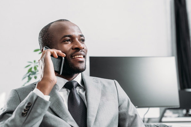 portrait of smiling african american businessman talking on smartphone in office - Фото, изображение