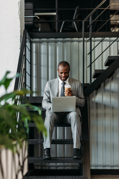 uomo d'affari afroamericano con caffè per andare con il computer portatile mentre seduto sulle scale
  - Foto, immagini