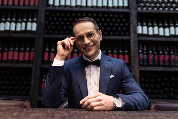 happy handsome man in stylish suit sitting in front of wine storage - Foto, Bild