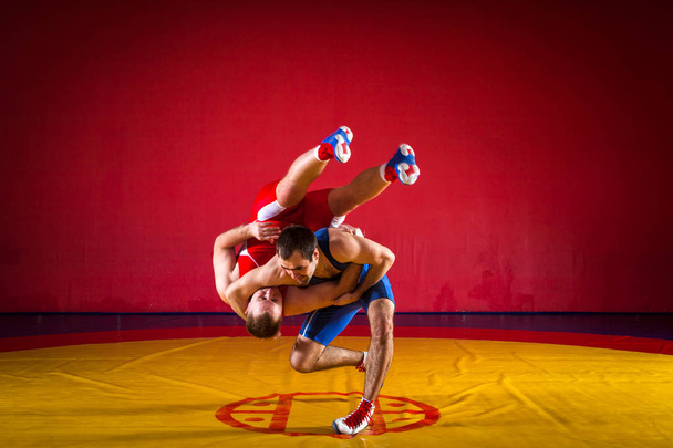 青と赤のレスリングのタイツを着た2人の若い男性がレスリングしており、ジムで黄色のレスリングカーペットの上で絶妙なレスリングを作ります - 写真・画像