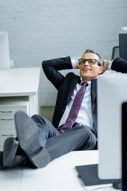 smiling businessman with hands behind head resting at workplace - Foto, Imagem