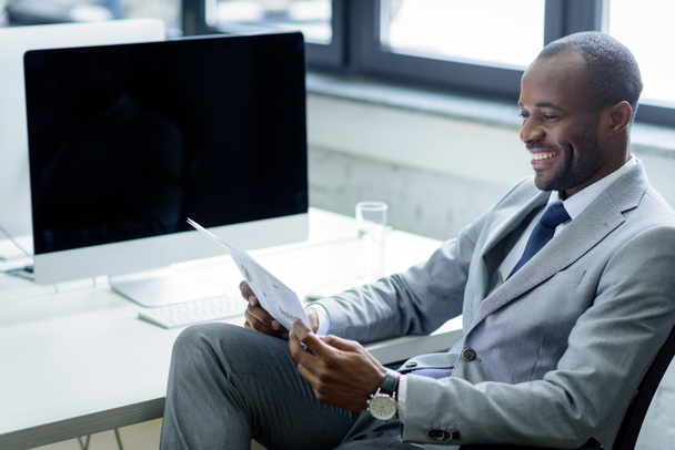 smiling african american businessman reading newspaper at workplace - Zdjęcie, obraz