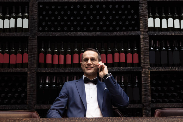 handsome man in stylish suit talking by phone at restaurant in front of wine storage shelves - Photo, Image