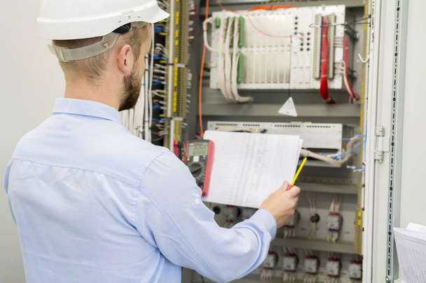 Engineer in white helmet reads design drawing against electric industrial panel. Service worker analyzes the electrical circuit in electrical automation control box with controllers. - Foto, Imagem