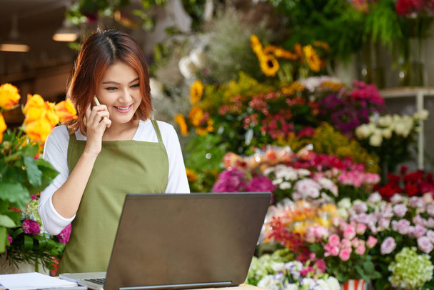 Přátelské vietnamské květina majitel obchodu na sobě zástěru mluví klientovi na smartphone stoje u přepážky, pas-stránky na výšku - Fotografie, Obrázek