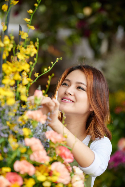 Getalenteerde jonge bloemist met charmante glimlach maken van kleurrijke bloemboeket terwijl verpakt in werk bij bloemenwinkel, portret shot - Foto, afbeelding