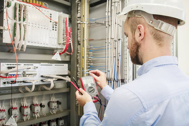 Service man with multimeter in hands adjusts electric control panel. Engineer in helmet tests device of protection. Worker checks electrical industrial equipment. Engineering work in automation box. - Zdjęcie, obraz