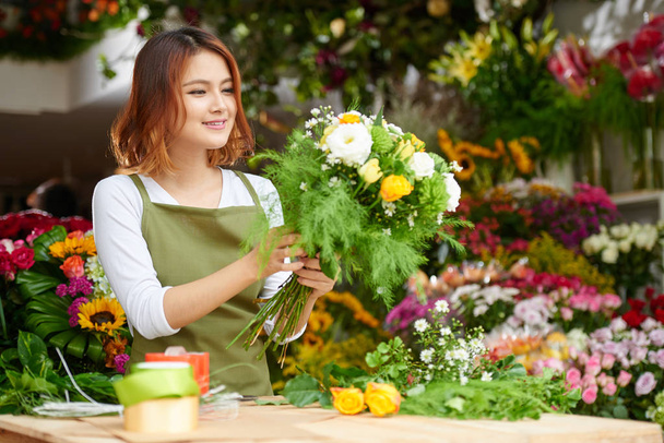 Азиатский флорист в фартуке стоит у стойки и делает букет для клиента, интерьер цветочного магазина на заднем плане
 - Фото, изображение