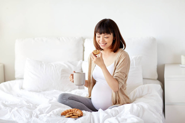 happy pregnant woman eating cookie in bed at home - Foto, Bild