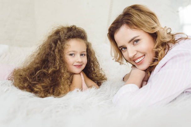 smiling mother and daughter lying on sheep skin blanket and looking at camera - Фото, изображение