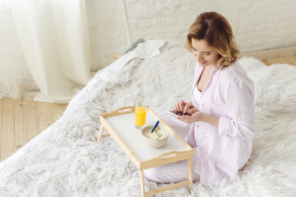 beautiful woman in pajamas using smartphone while sitting on bed with breakfast on tray - Foto, afbeelding
