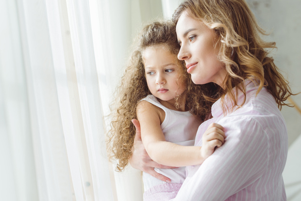 beautiful mother looking at window and holding on arms her curly daughter  - Photo, Image