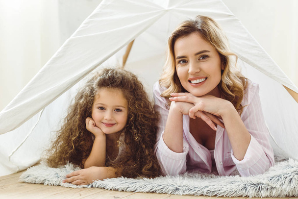 smiling mom and happy curly daughter lying in kid wigwam - Foto, afbeelding
