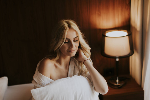 Портрет счастливой блондинки, обнимающей подушку дома в спальне
 - Фото, изображение