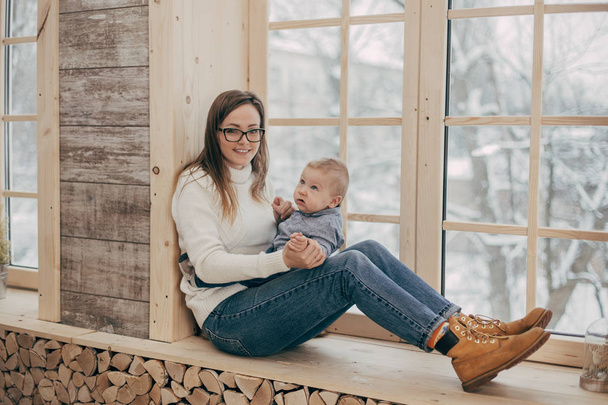 Portret van lachende jonge vrouw met babyjongen zitten op de vensterbank - Foto, afbeelding
