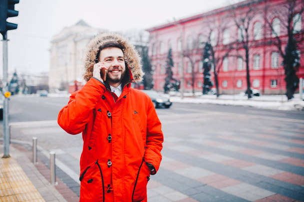 young guy with beard and red jacket in hood a student uses mobile phone, holds in his hand near the head, talking on the phone with smile on the background of red building the university or college - Photo, Image