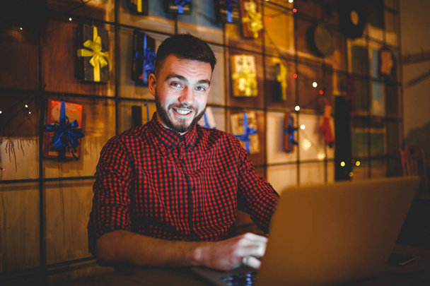 Formation thématique et ordinateurs. Un jeune homme avec une barbe et une chemise utilise un ordinateur portable, des impressions sur le clavier dans un café à une table en bois le soir. Décor de Noël et accrocher une guirlande
 - Photo, image