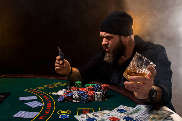 Uomo barbuto con sigaro e vetro seduto al tavolo da poker in un casinò. Gioco d'azzardo, carte e roulette
. - Foto, immagini