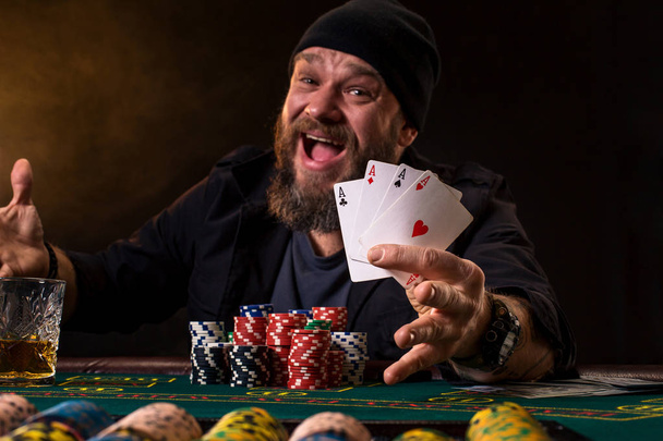 Bearded man with cigar and glass sitting at poker table and screaming isolated on black - Photo, Image