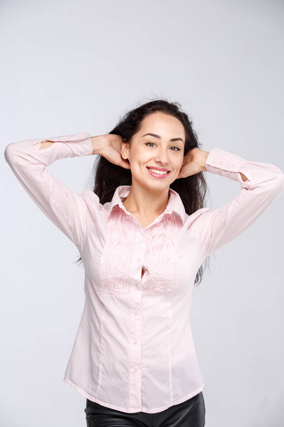 Close-up portrait of a young very beautiful woman with a charming toothy smile, black hair and brown eyes on a white background in a pink shirt. Hands near the head. Positive and joyful emotions - Photo, Image