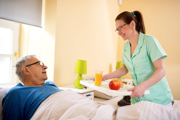 Young smiling nurse brings breakfast to senior patient - Photo, Image