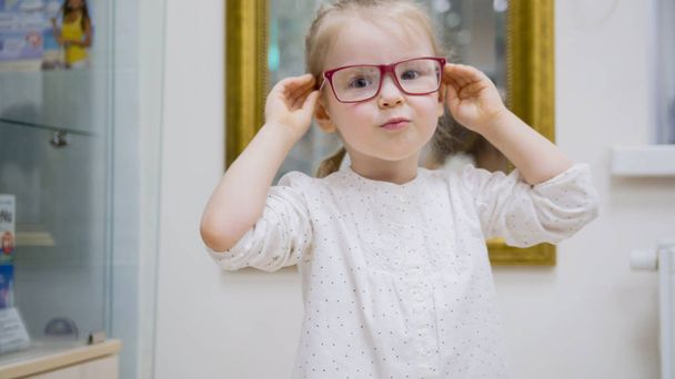 Маленькая девочка пробует модные медицинские очки возле зеркала - шоппинг в офтальмологической клинике
 - Фото, изображение