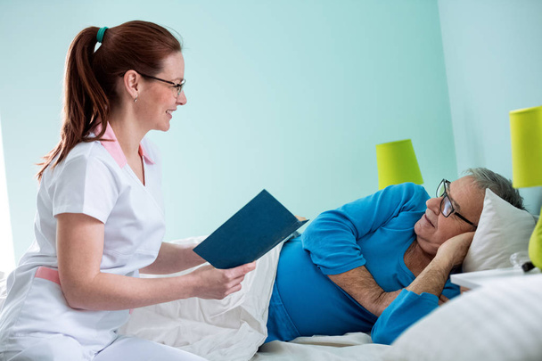 Smiling positive nurse reading a book to her old patient - Foto, immagini
